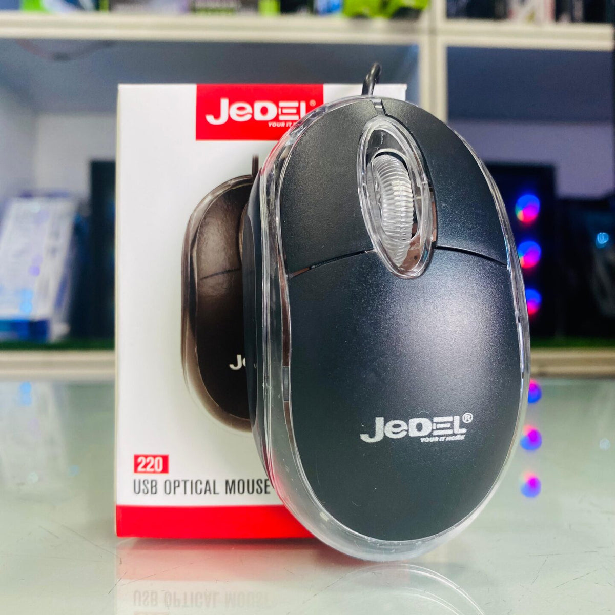 Jedel 220 USB optical mouse unboxed black mouse leaning against original box in tech store with RGB gaming PCs in background